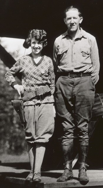 Rangers Eva and Charles McNally stand on the porch of the ranger station.  She is wearing a badge and a gun holstered on her hip.