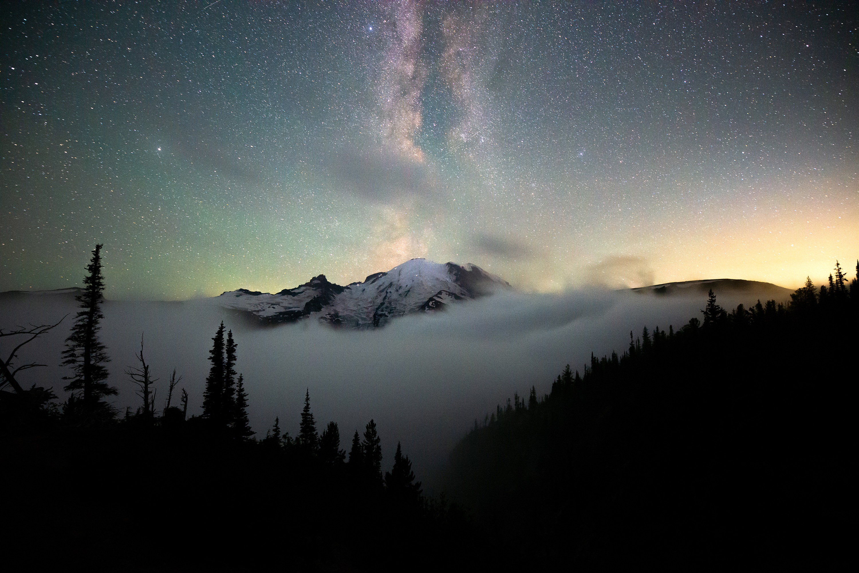 Nighttime picture of Milky Way galaxy above a snow-covered mountain and clouds as seen through silhouetted evergreens.
