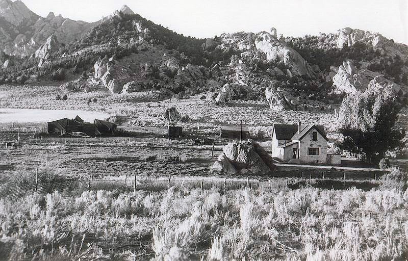 Tracy House Rustic stone farmhouse sits in field surrounded by arid landscape and rocky mountains.