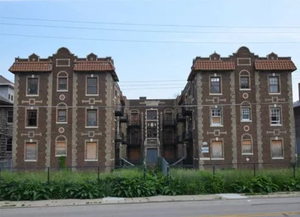 The Ambassador apartment building Image of a three-story brown brick apartment building with two symmetrical facades in Mediterranean Revival style. Some windows are boarded up. Overgrown greenery separates the building and a fence.