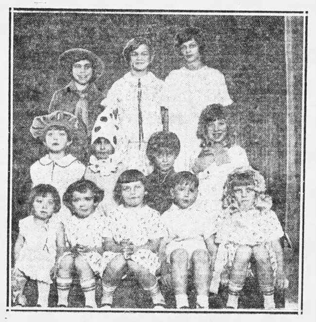 Black and white photo of school children arranged in three rows. Some wear costumes and hats.