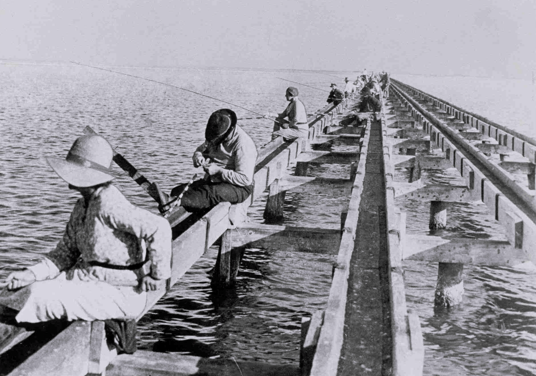 A black and white photo of people fishing off a pier.