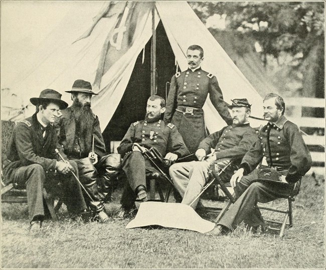 Black and white image of six men, 5 seated and one standing, in civil war uniforms in front of a canvas tent.