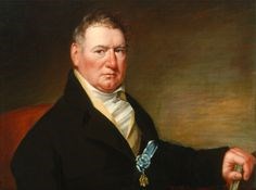 Portrait of man, receding hairline, dark coat, white shirt, blue medallion