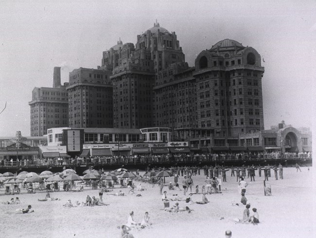 Monumental hospital building looms over boardwalk and people recreating at a beach