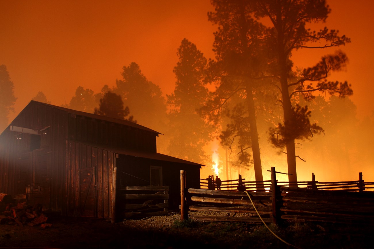 Fire burns near wooden barn at night with firefighters providing protection.
