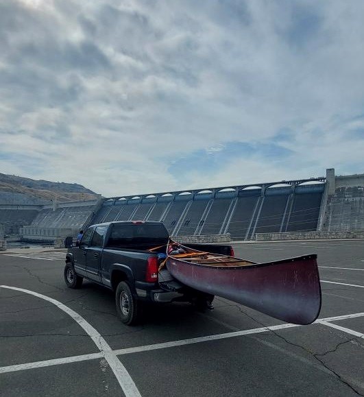 Transporting their canoe past the Grand Coulee Dam. A dark blue truck holds a red canoe in the truck bed. It is parked in a parking lot overlooking the Grand Coulee Dam.