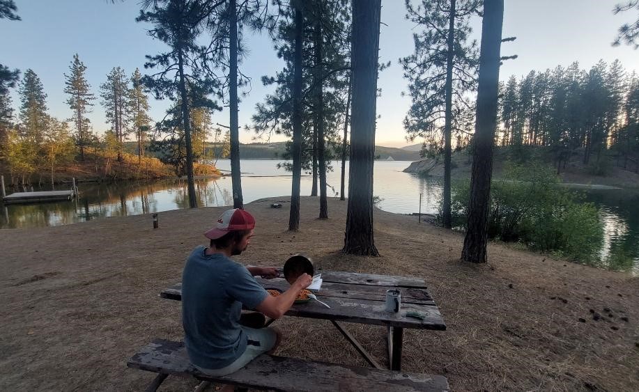 Sitting down for dinner along Lake Roosevelt. A man sits at a wooden picnic bench, eating a cooked meal, with trees surrounding a quaint campground on the lake.
