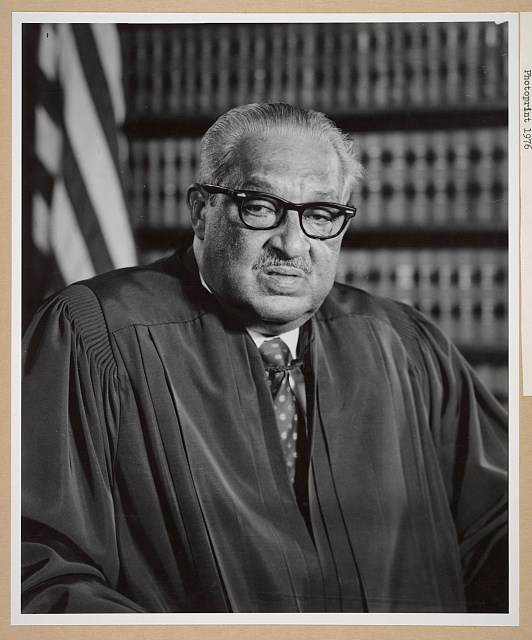 Black and white portrait of an older African American man wearing judicial robes and glasses, seated in front of a wall of legal books with a U.S. flag partially visible in the background.