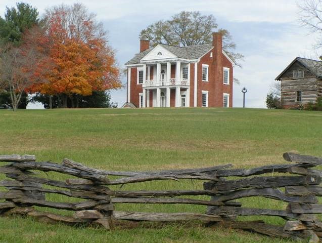 A brick mansion sits on top of a hill with a log cabin next to it.