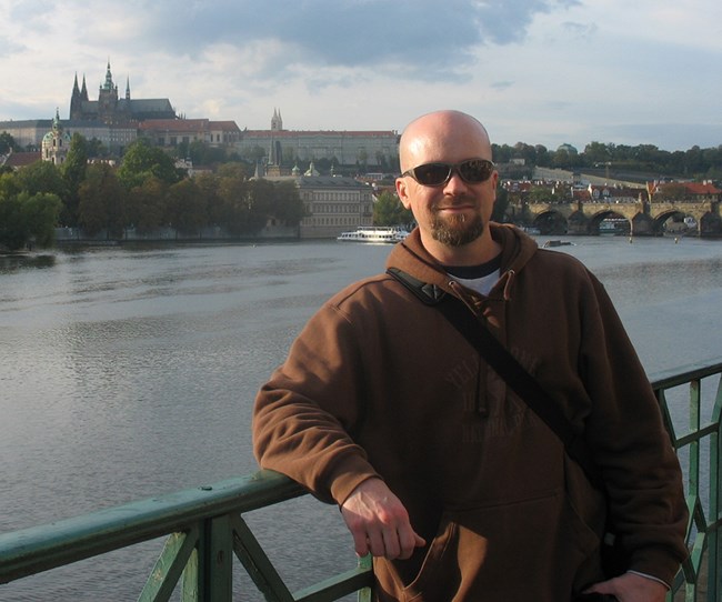 A man stands on a bridge (in Prague) with a castle in the background.