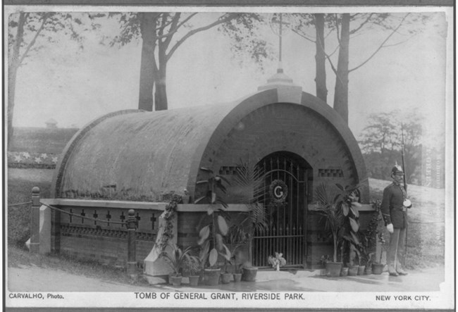 Black and white photo of a man standing on guard in a military uniform in front of a small black temporary tomb structure