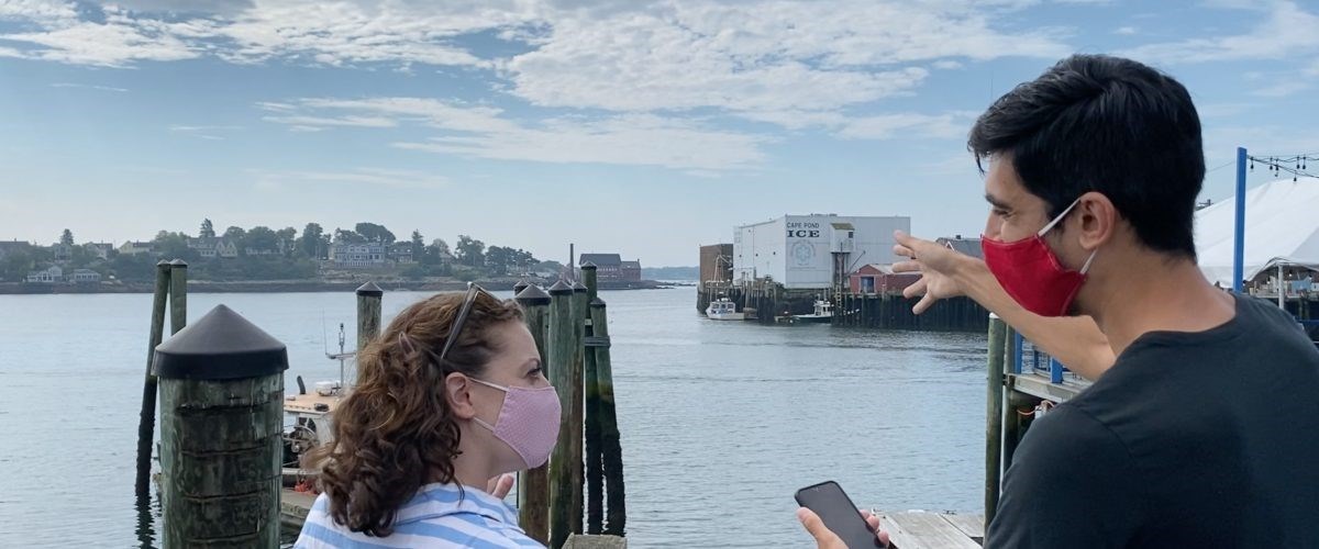 Trails and Sails Two people wearing surgical masks stand along harbor with ocean view and inlet coast of town across water. One holds phone and points arm.