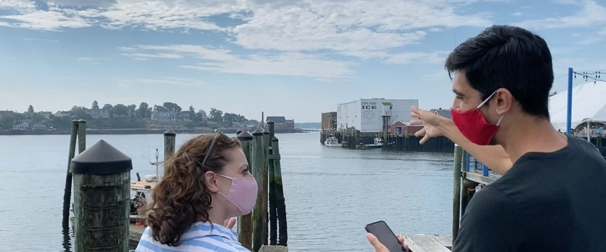 Trails and Sails Two people wearing surgical masks stand along harbor with ocean view and inlet coast of town across water. One holds phone and points arm.