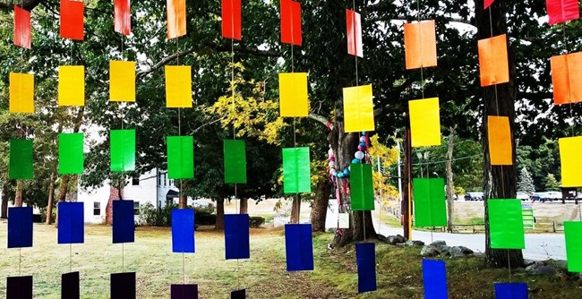 Trails & Sails Abstract sculptural work made of an array of rainbow rectangular tiles connected by string or wire. Green grass of park in background.