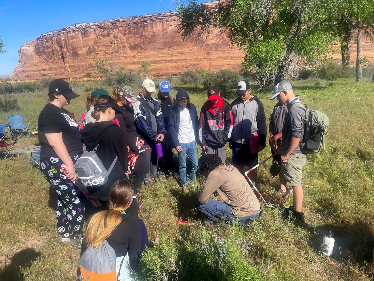 Training young citizen scientists Group of standing students surround a woman sitting on the ground in a grassy field, with measuring equipment. Red rock cliffs surround them.
