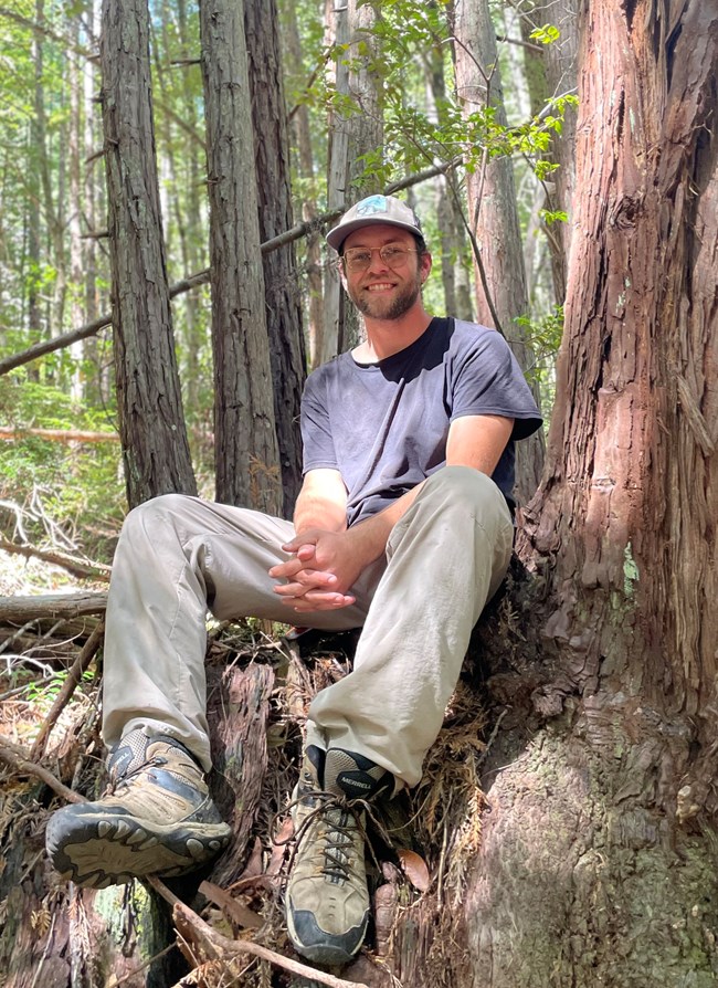 Young man sits at the base of a tree in the forest.