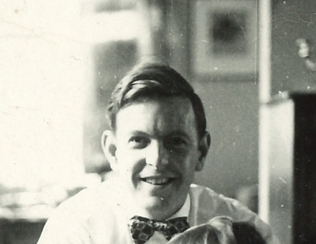 Smiling black-and-white portrait of a youthful man in a white shirt and bow-tie with a blurry interior background.
