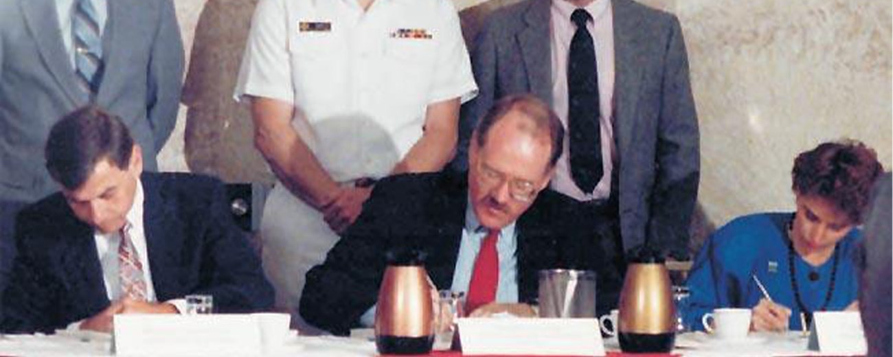 Signing the Tri-Party agreement Two men and one woman sign documents at a table while three men stand behind.