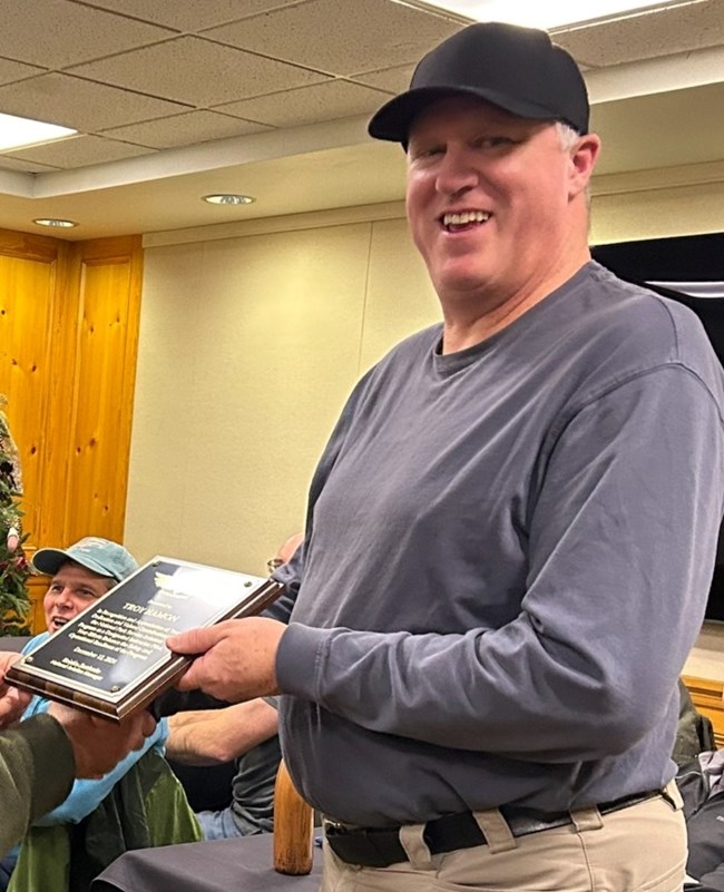 A man with a dark baseball cap holds a plaque and smiles at the camera.