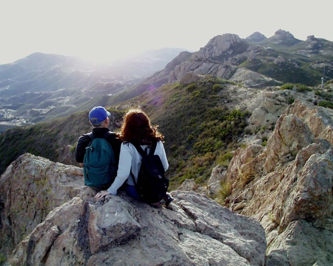 Two individuals sitting atop a rocky outcrop while watching the sun set