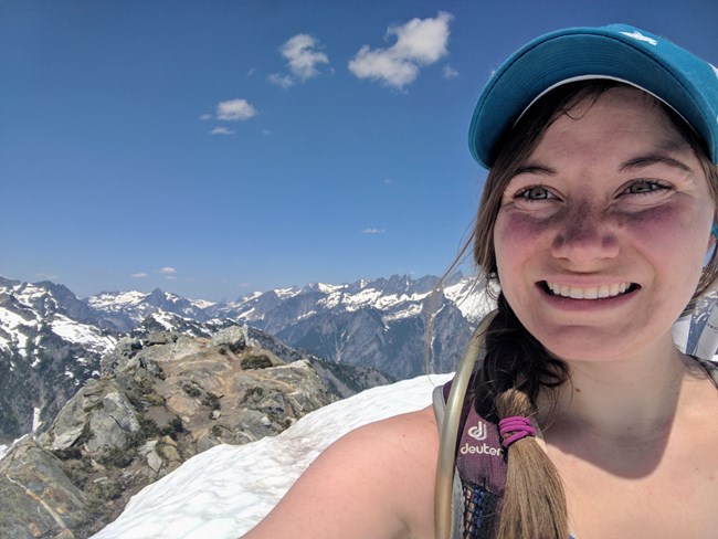a selfie of smiling young woman in a baseball hat with mountainous view in the background