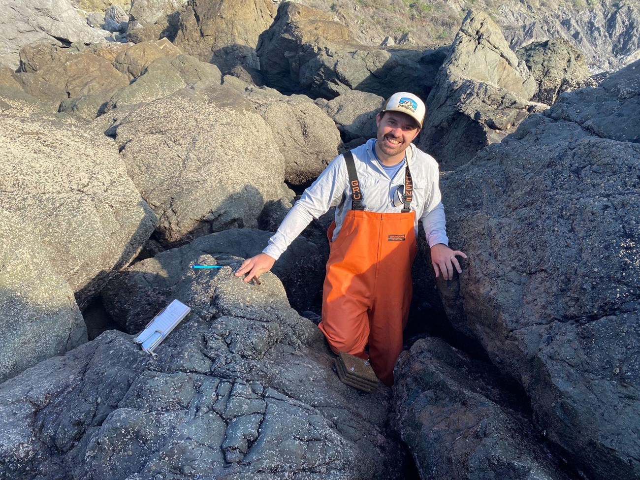 A person wearing orange waterproof overalls smiles at the camera while standing between two rocks in the intertidal zone. A clipboard and pencil are off to the side.