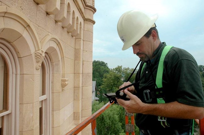 A worker with a harness and hardhat on a lift beside a sandstone building with arched windows enters data into a tablet