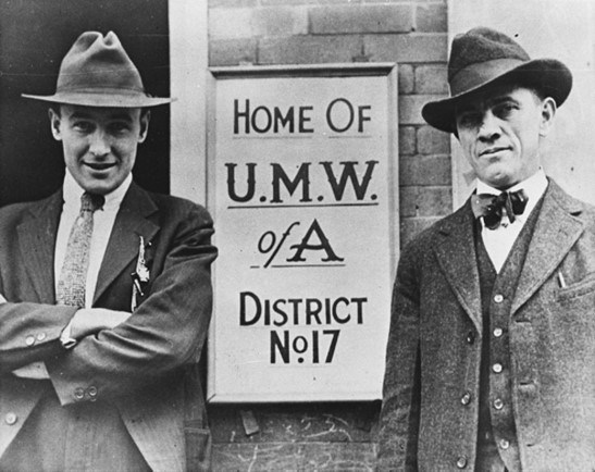 Two smiling men in suits and hats stand beside each other with a sign in between them reading Home of U.M.W. of A. District No. 17.