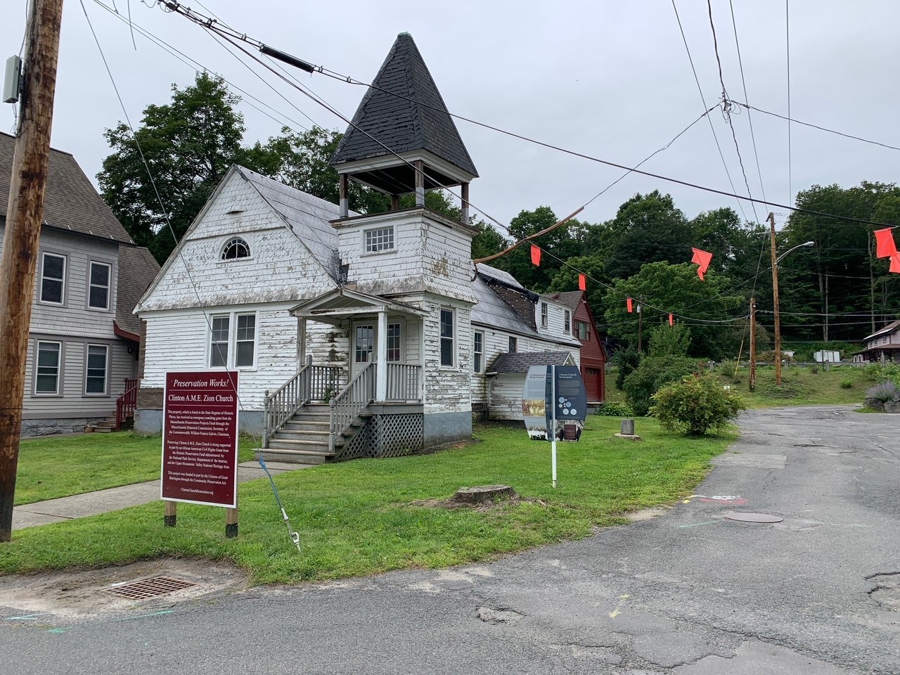 Clinton Church Restoration A church building with weathered white shingle siding and large turret above entrance. Green grass and historical marker sign in front, asphalt road at right.