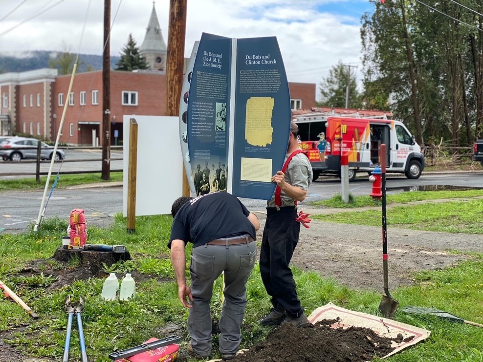 Clinton Church Restoration Two people hoist and plant interpretive sign on lawn. The sign includes four display panels set 90 degrees apart like a plus sign, creating eight informational sections about the church, W.E.B. DuBois and the A.M.E. Zion Society