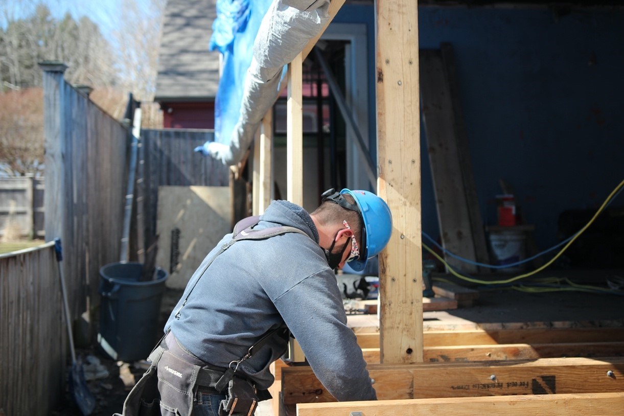 UPHV6 Construction crew member wearing hard hat and mask works on exposed beam section of wall of building