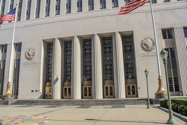 Front of stone government building. A flight of low steps leads to 4 sets of gold double doors separated by Greek columns with decorative metalwork in between. “United States Courthouse” seals featuring eagles and large American flags flank the entrance.