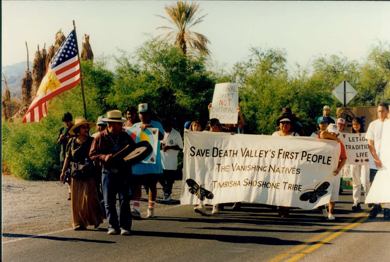 Timbisha Shoshone members and allies marching on Memorial Day 1996 to demand restoration of their homeland. Group marching in street with sign stating "Save Death Valley's First People"