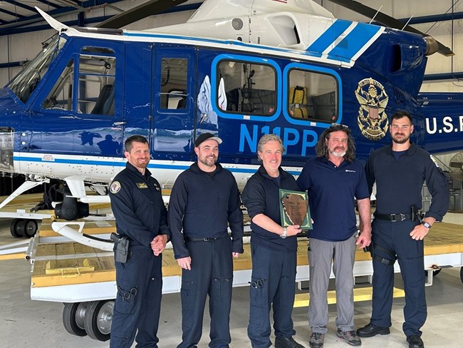 Five men, middle one holding plaque, stand in front of blue helicopter.