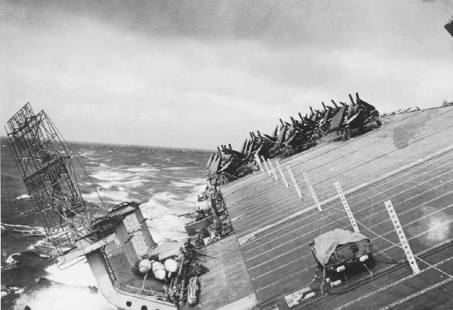 Black and white photograph of an aircraft carrier tipping its deck into the ocean