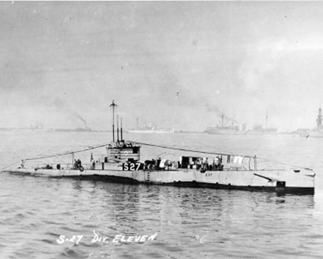 submarine with S-27 painted on it above the water, with other ships in background.