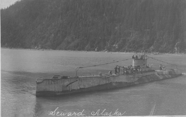 Submarine with S-32 painted on it, above water, with forested shoreline in background. Text says "Seward, Alaska"