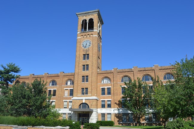 Multi-story tan brick factory building with a central clock tower. The main entrance is located at the base of the clock tower. On either side of the tower, there are several sets of windows on each floor. The windows on the top level are arched.