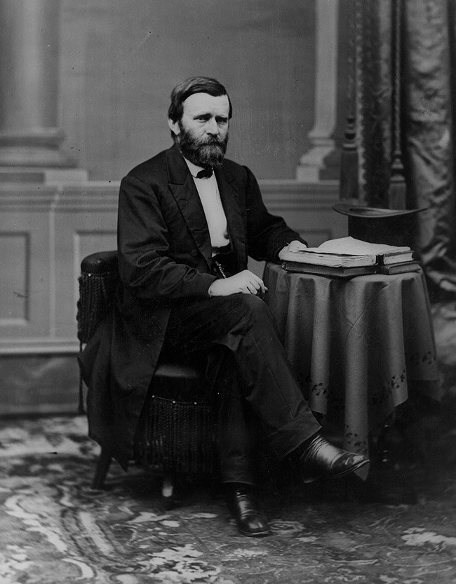 Black and white image of a man in a suit facing right sitting in a chair at a desk