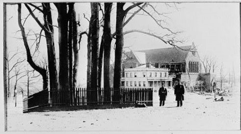 Photograph of a grove of trees surrounded by picket fence, with two uniformed officers in circa 1900 standing next to the fence.