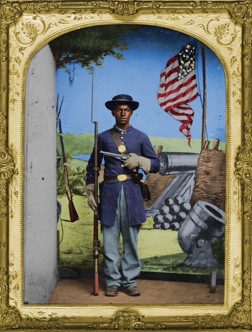 Colorized photo of an African American soldier during the Civil Wear wear coat, pants, and hat while holding two weapons.