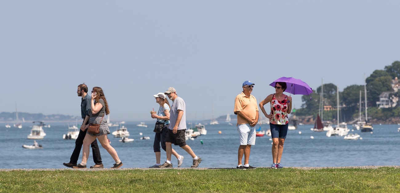 People enjoying a sunny day Groups of people walking on path with water and boats in the background.