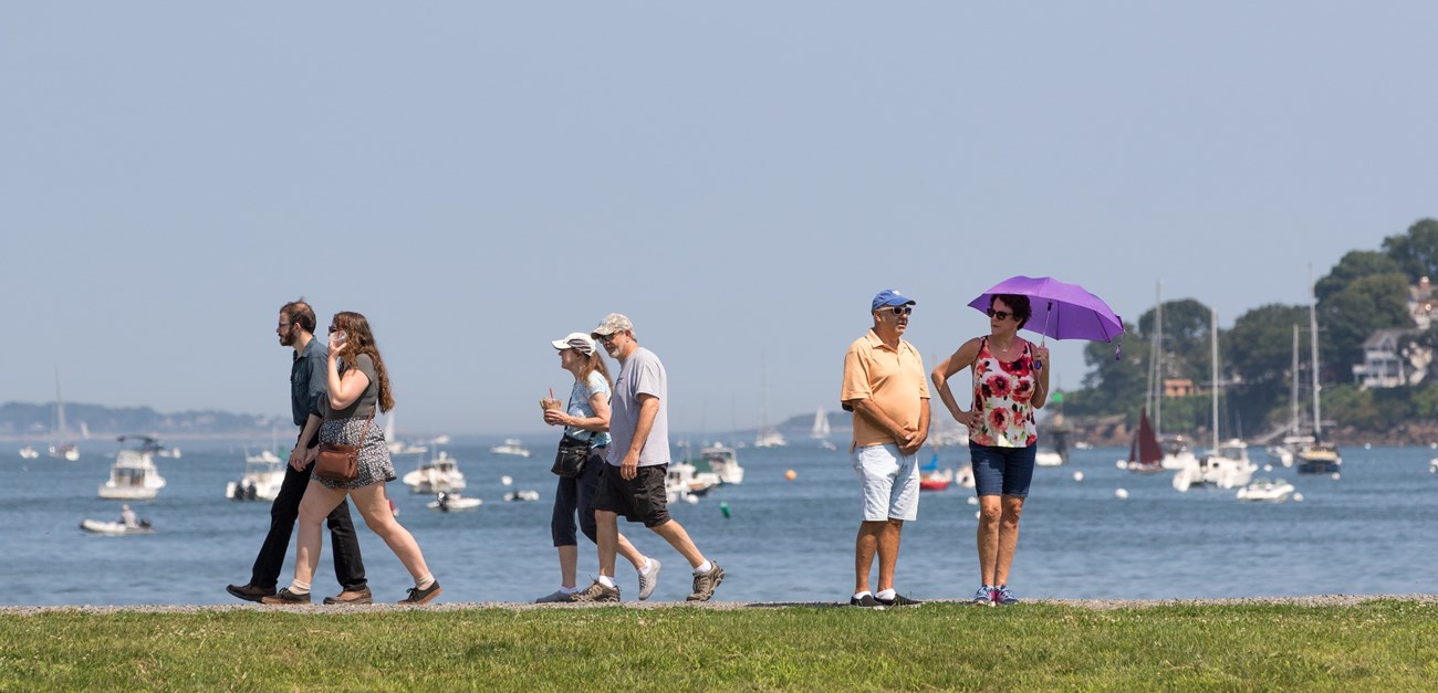 Groups of people walking on path with water and boats in the background.