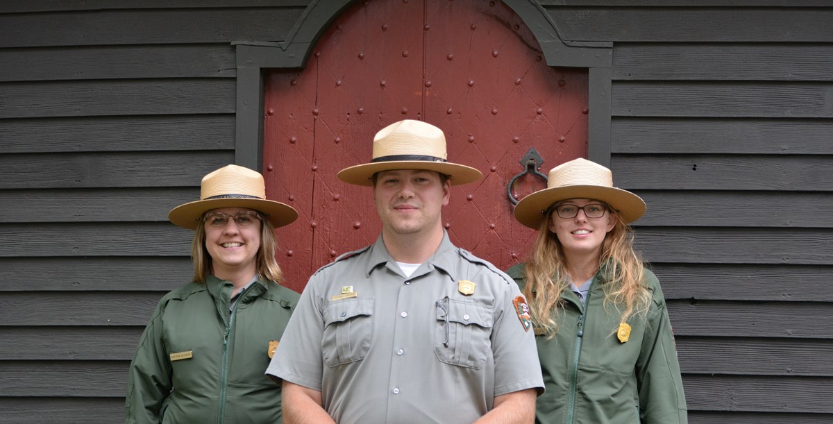 Three smiling rangers in tan flat hats stand in front of a red door.
