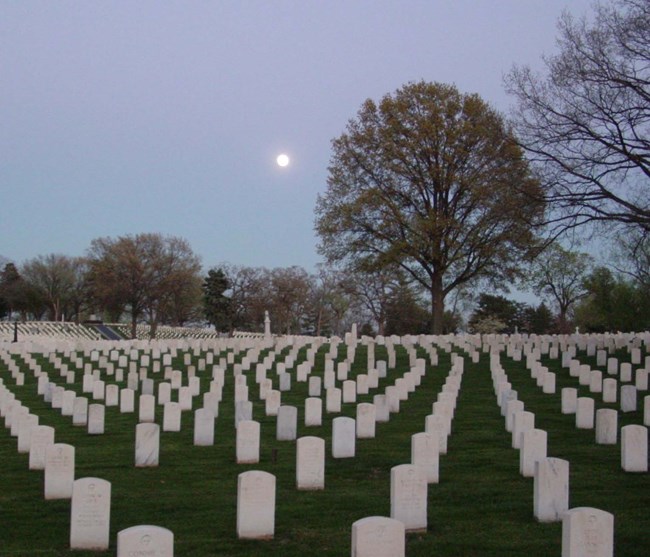 Cemetery with rows of white grave markers stretching into rolling fields, landscaped grass and trees.