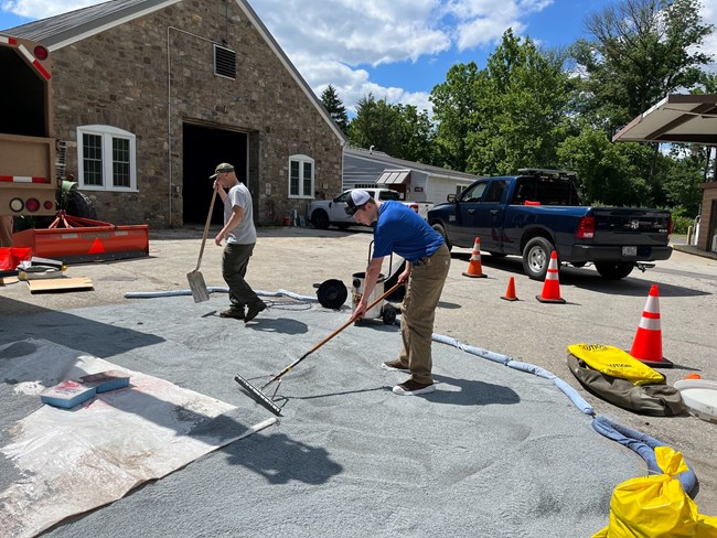 Two men work with tools moving gravel over pavement near a building.