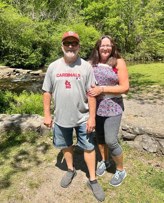 A man wearing a gray tshirt, baseball cap and glasses poses next to a woman with brown hair, floral shirt and capri pants.