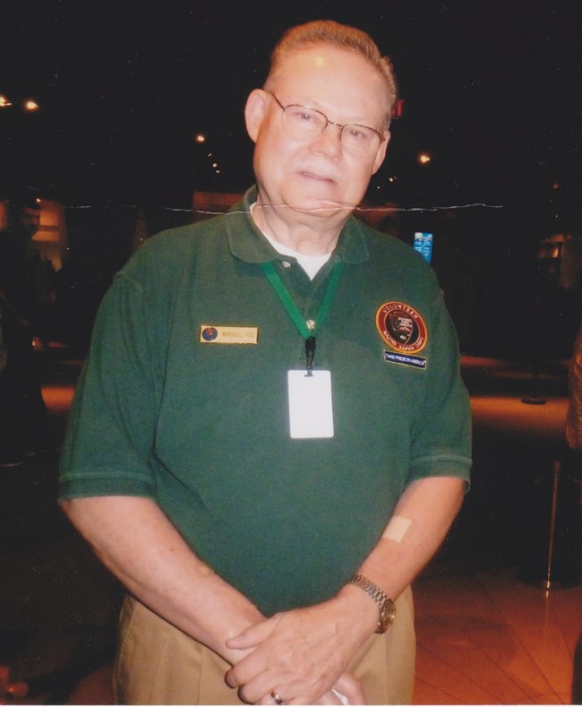 Portrait of a man from the waist up in a dark room. He is looking at the camera and is wearing a green volunteer polo shirt.