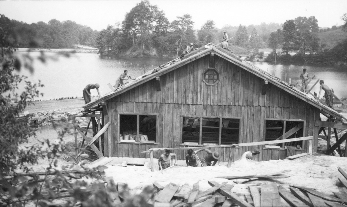Black and white photo of a wooden building under construction; shirtless men dig in the foreground and work on the roof; in the background, a large body of water and trees.